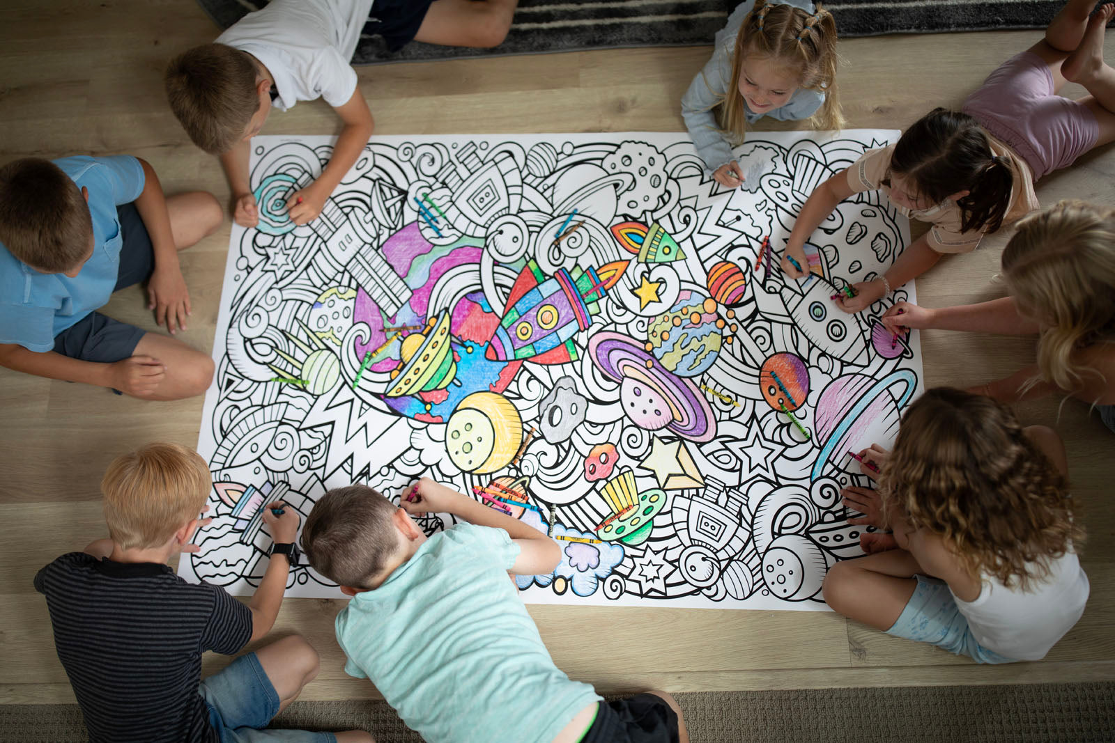 A group of children sitting around a large coloring pad on a wooden floor, coloring a colorful space-themed cartoon design with rockets, planets, and astronauts.
