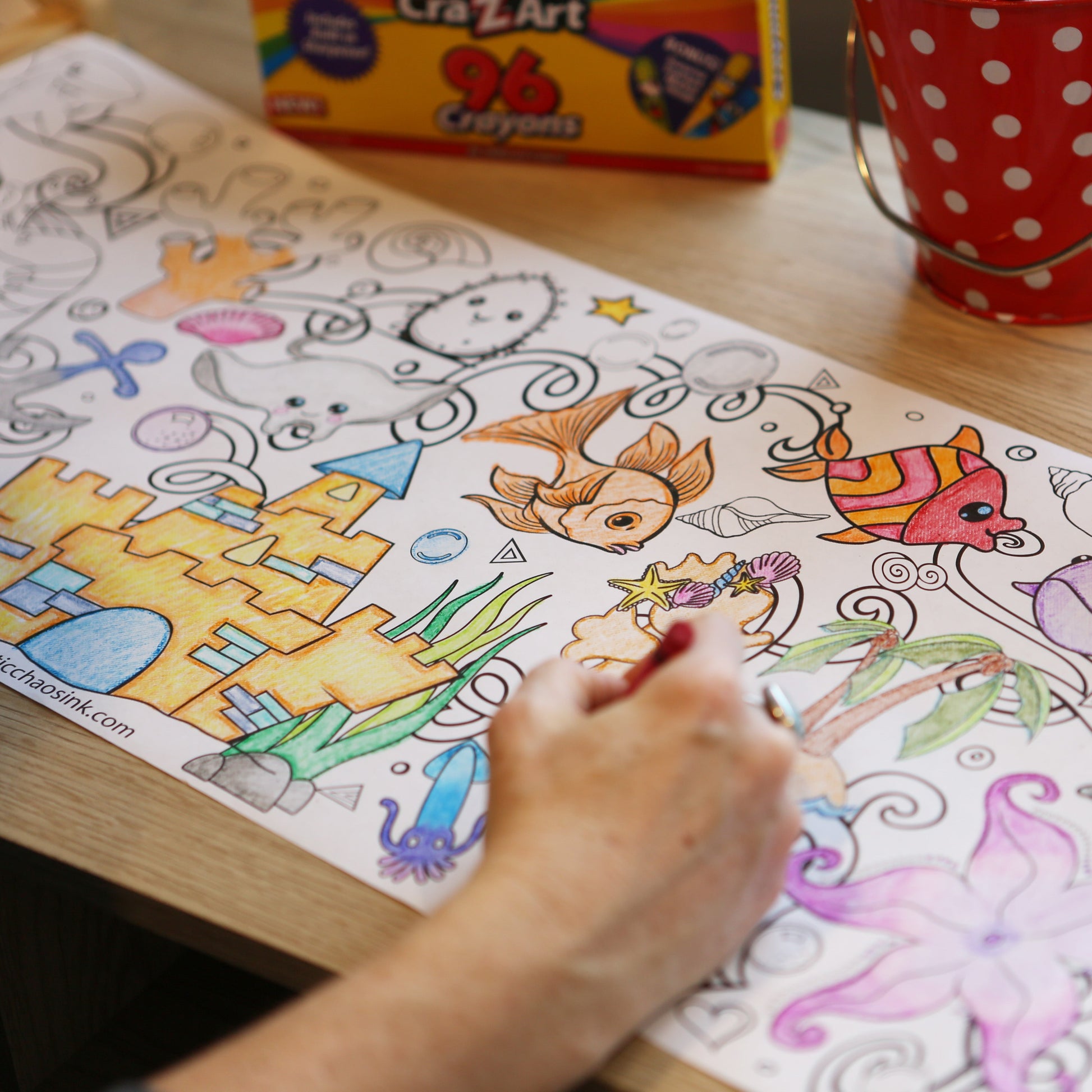 Coloring book with underwater scene on a table, hand holding crayon.