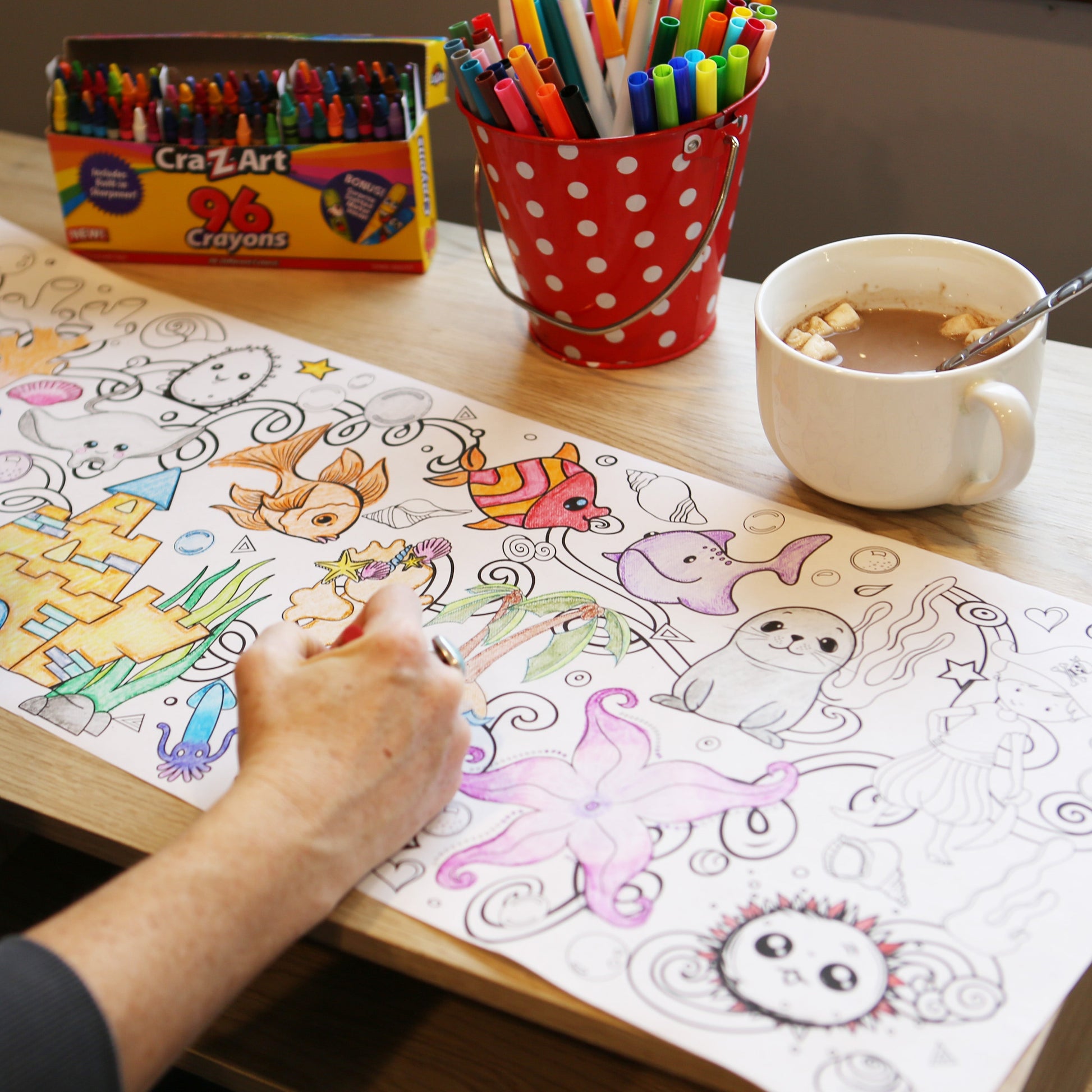 Person coloring an ocean picture with markers on a table next to a mug of hot chocolate and a box of CrazArt crayons.